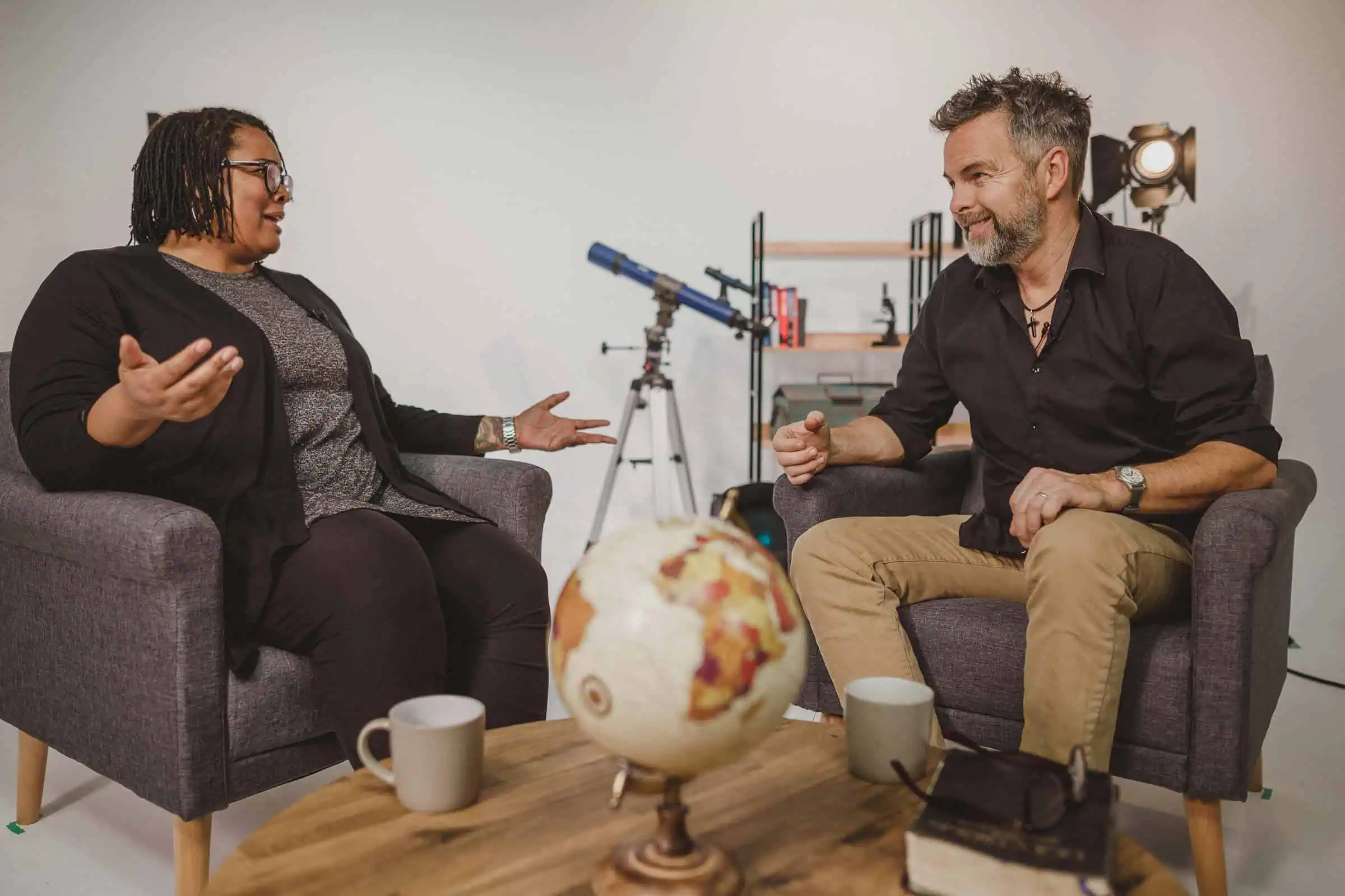 Een vrouw en een man zitten in twee fauteuils in gesprek, met een wereldbol op de houten salontafel tussen hen in.