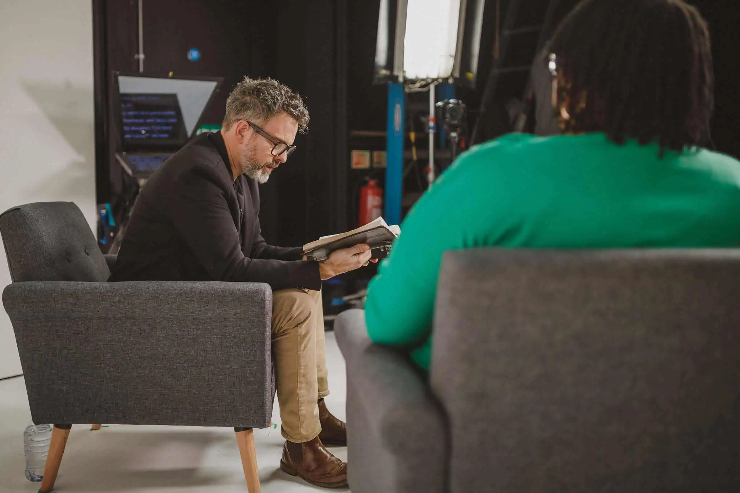 Man met bril leest uit een Bijbel in een studio-opstelling met een autocue, tegenover een vrouw in een groene trui die op de rug zichtbaar is.