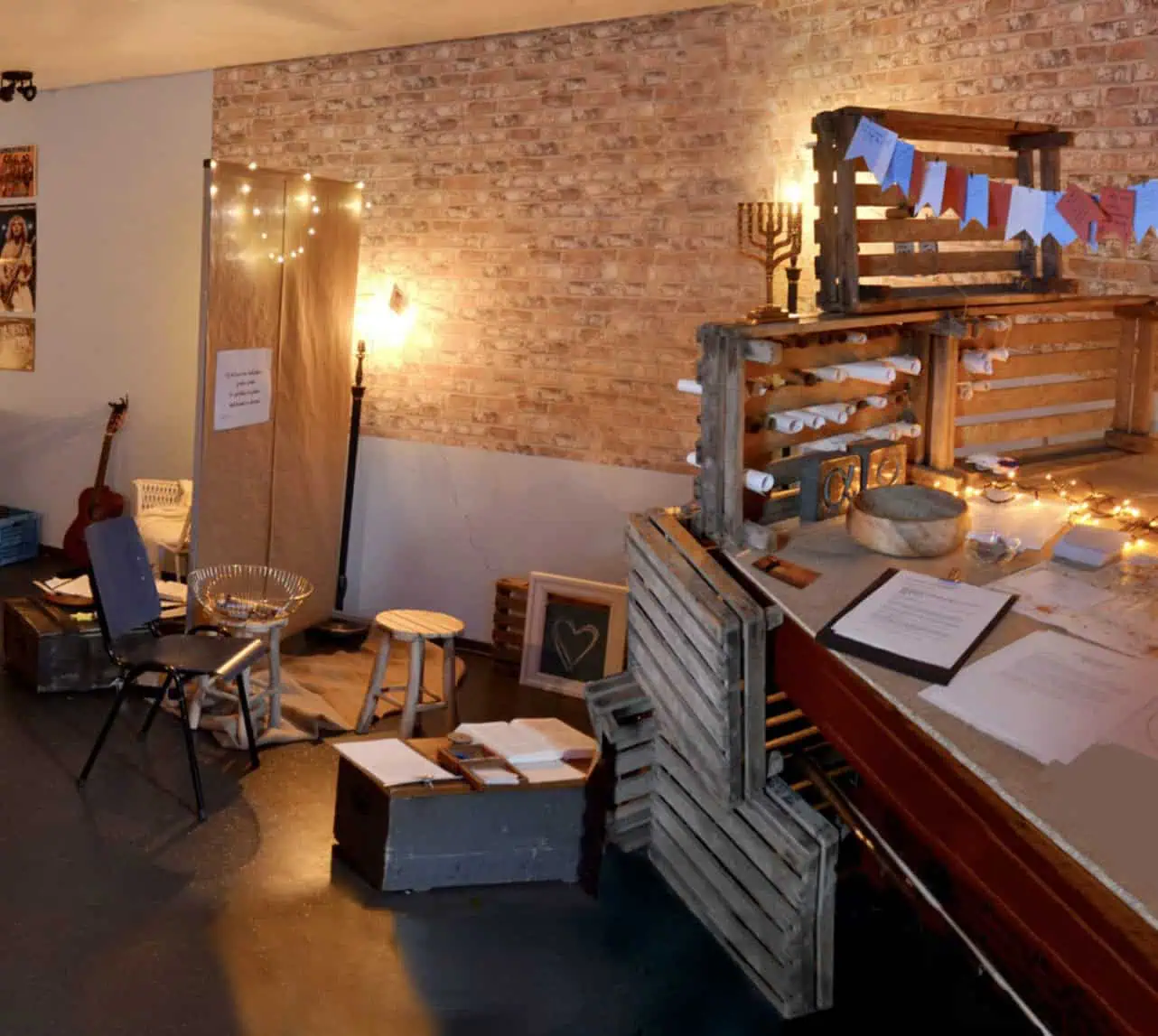 Sfeervolle interactieve gebedsruimte met een bakstenen muur, lichtsnoeren, houten kratten, een menora, een gitaar en handgeschreven gebedskaartjes.