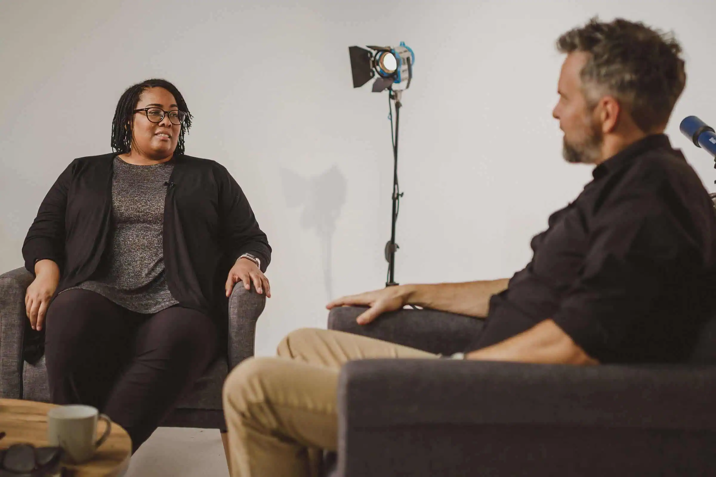 Een vrouw en een man zitten tegenover elkaar in gesprek in een studio, met een filmlamp op statief tussen hen in.
