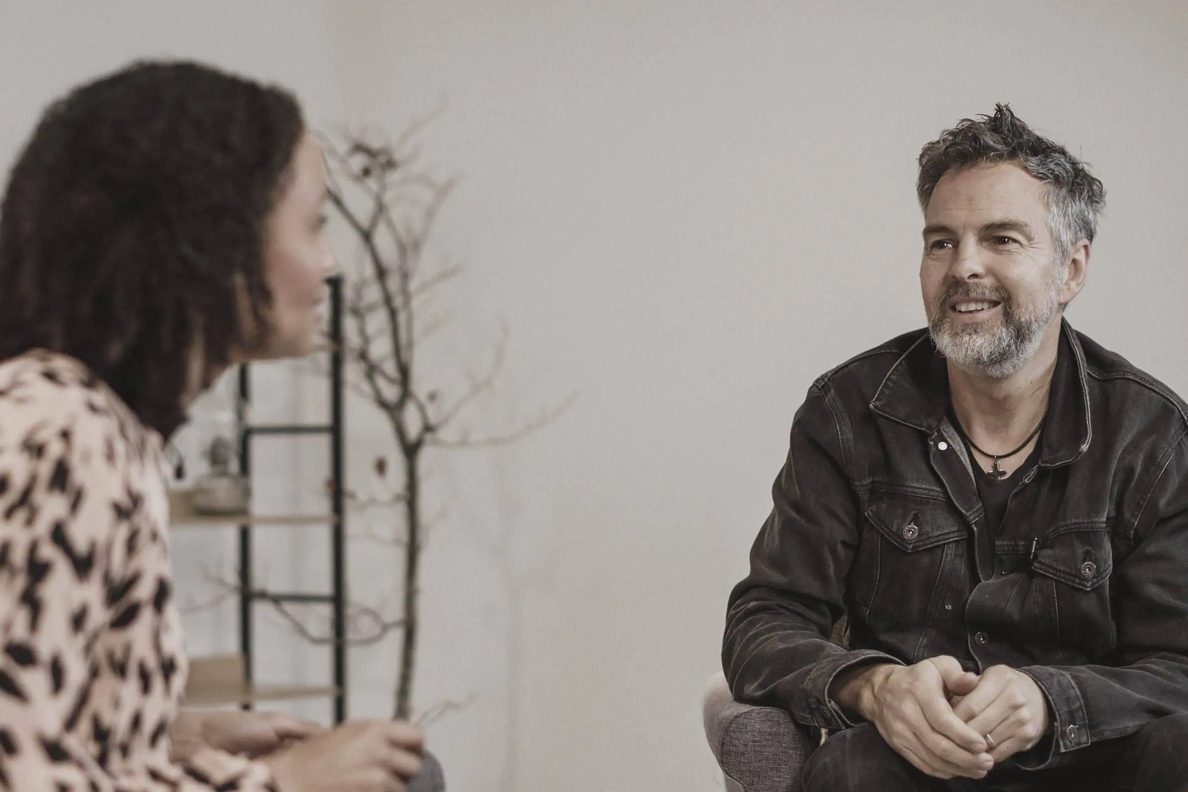 Man met grijze baard en spijkerjasje zit glimlachend met gevouwen handen in een fauteuil in gesprek met een vrouw die op de voorgrond onscherp zichtbaar is, met op de achtergrond een kale tak en een open kast.