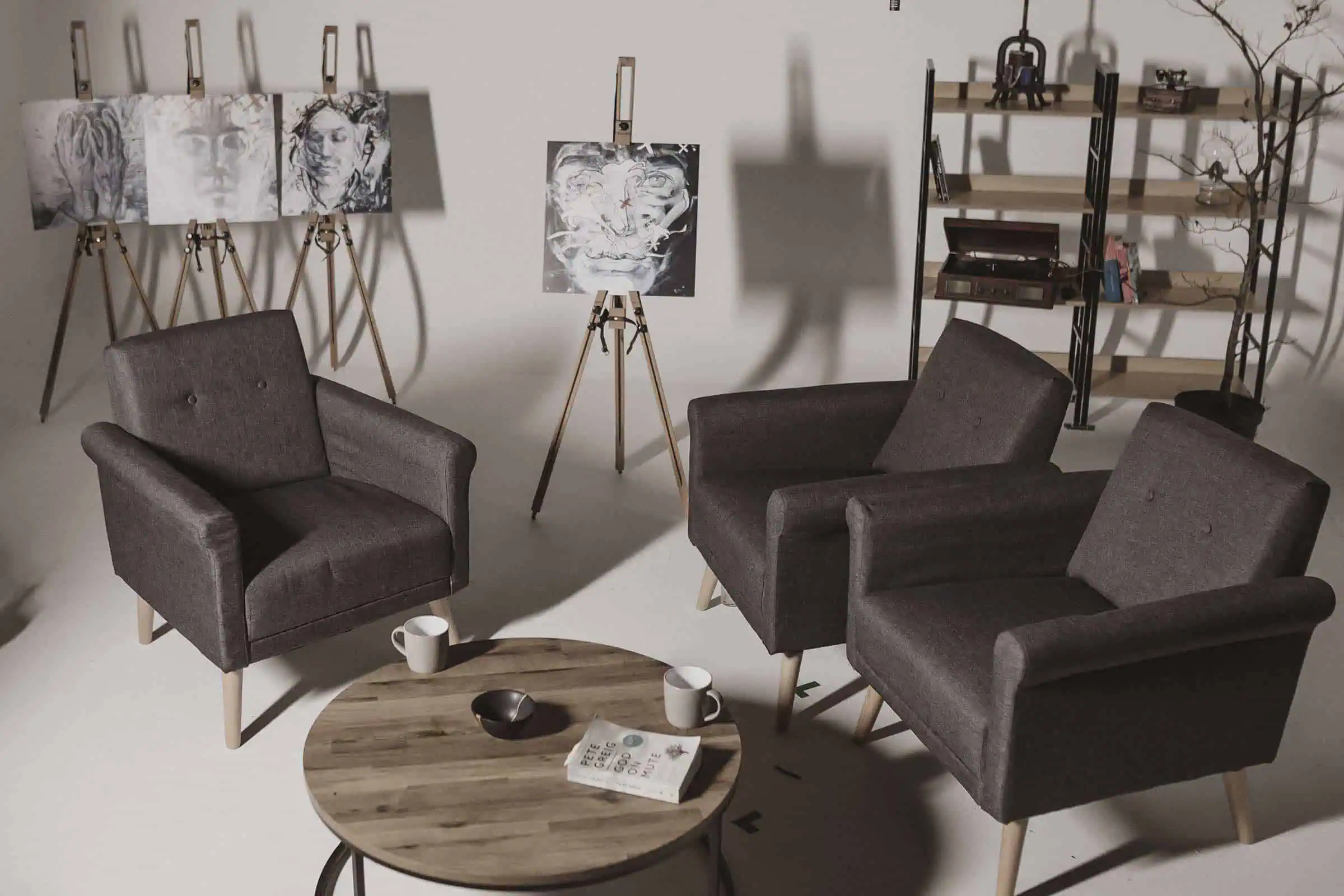 Studioset met drie grijze fauteuils rond een ronde houten tafel met mokken en een boek van Pete Greig, met houtskoolportretten op ezels en een boekenkast met platenspeler op de achtergrond.
