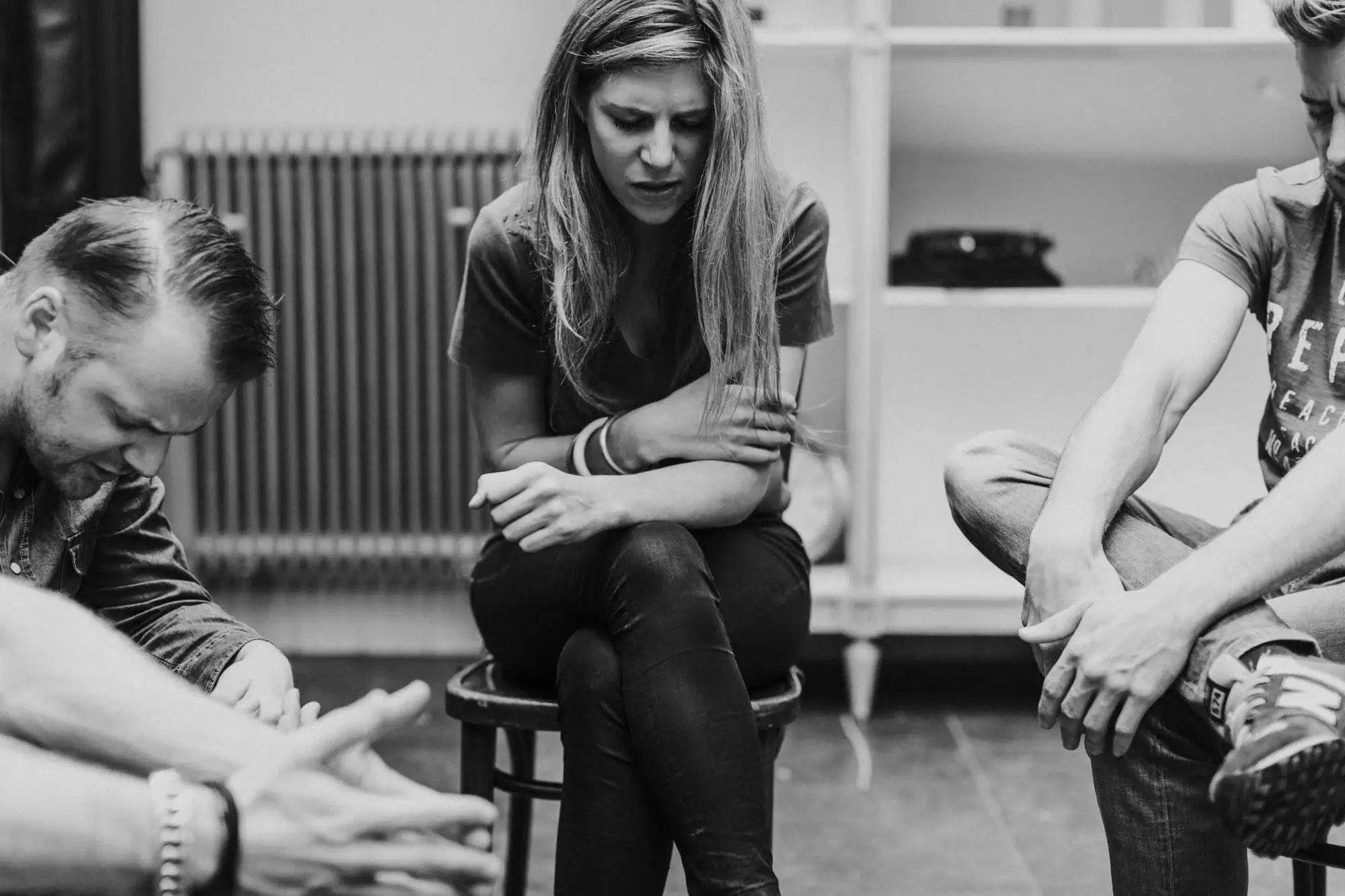 Zwart-witfoto van drie mensen die in een kring met gebogen hoofden zitten te bidden, met een vrouw op een kruk in het midden.