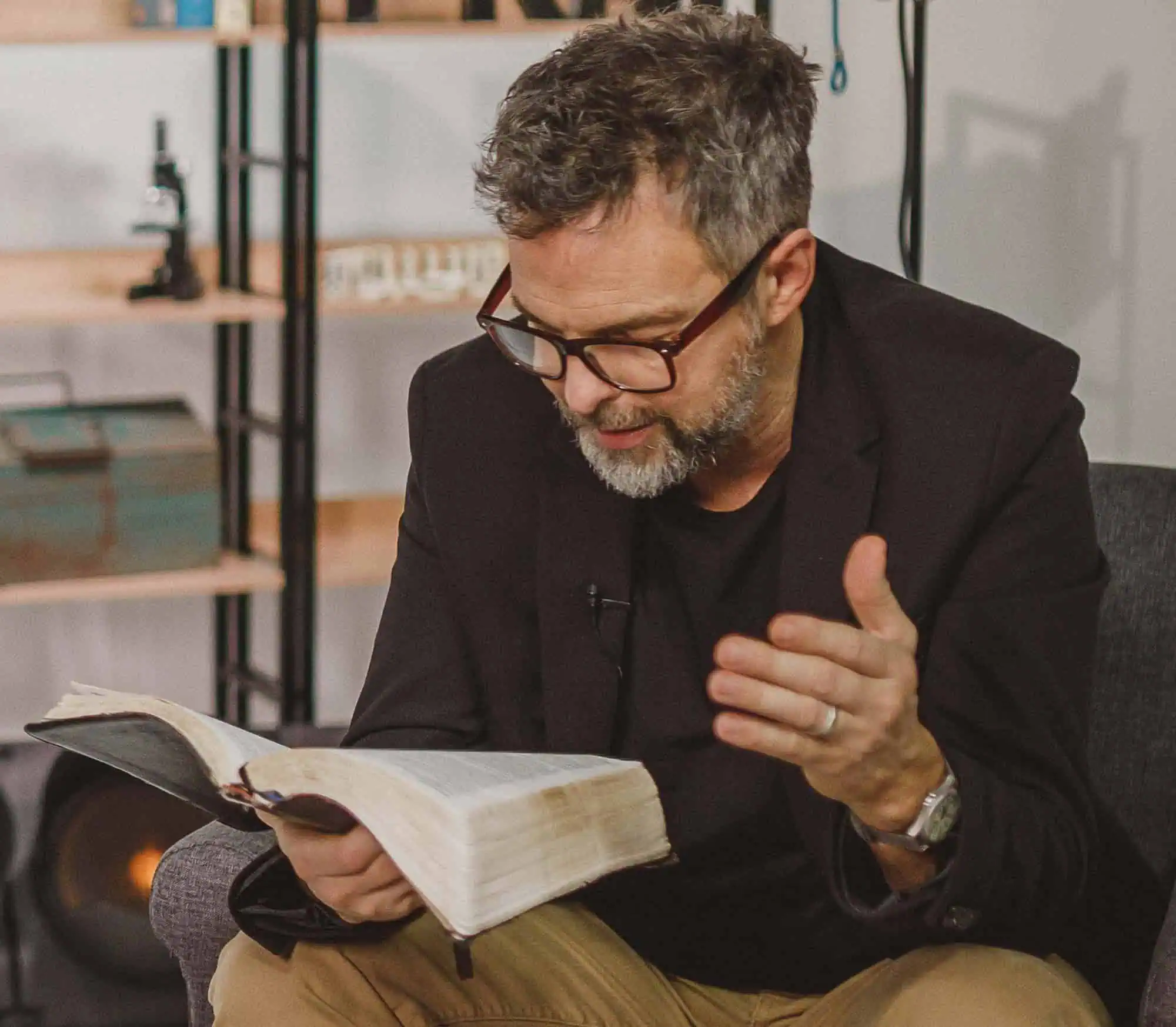 Man met bril en grijze baard leest uit een Bijbel en gebaart met een hand tijdens een sessie in een studio, met op de achtergrond een boekenkast met een microscoop