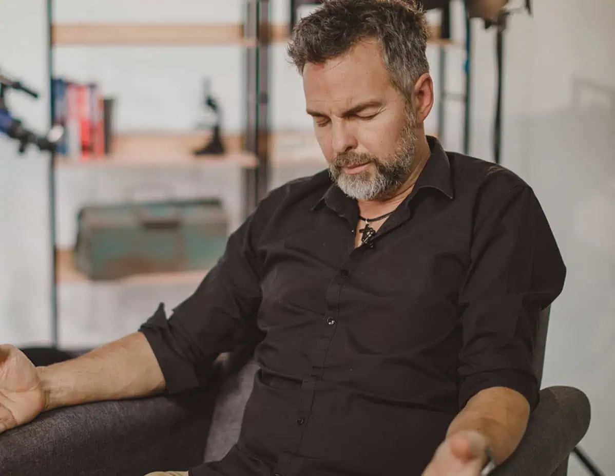 Man met grijze baard en zwart overhemd zit in een fauteuil met neergeslagen blik te praten, met op de achtergrond een boekenkast met een telescoop.