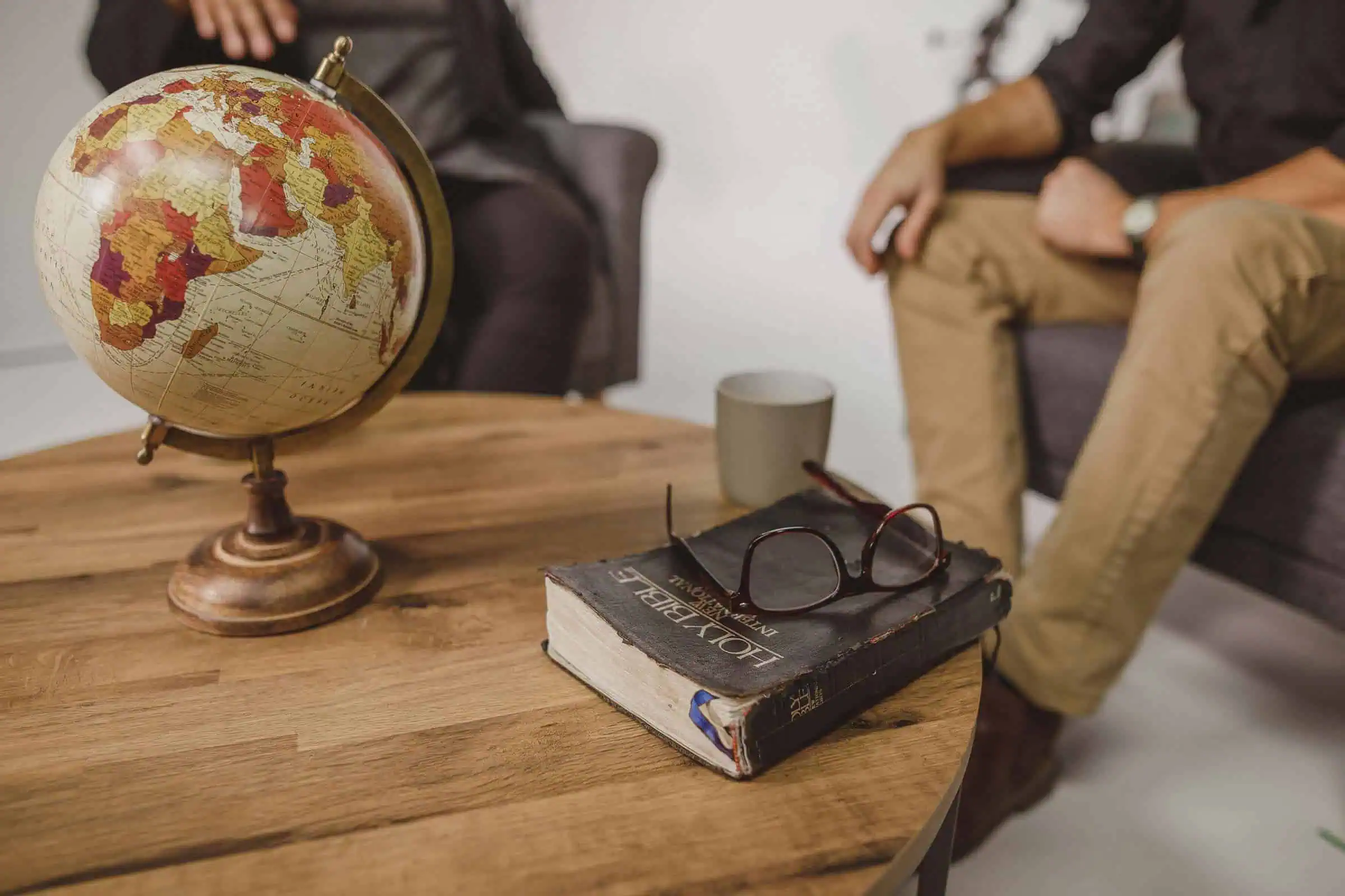 Een antieke wereldbol en een versleten Holy Bible met een leesbril erop, op een houten salontafel, met op de achtergrond twee zittende personen.