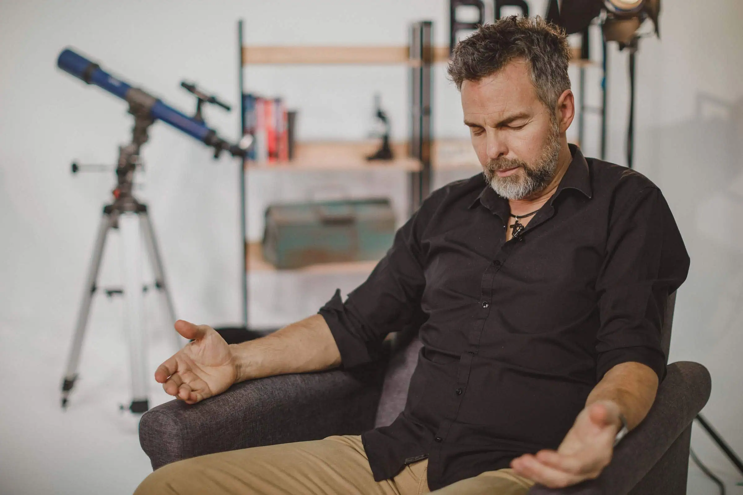 Man met grijze baard zit in een fauteuil met gesloten ogen en open handen te bidden, met een telescoop en boekenkast op de achtergrond.
